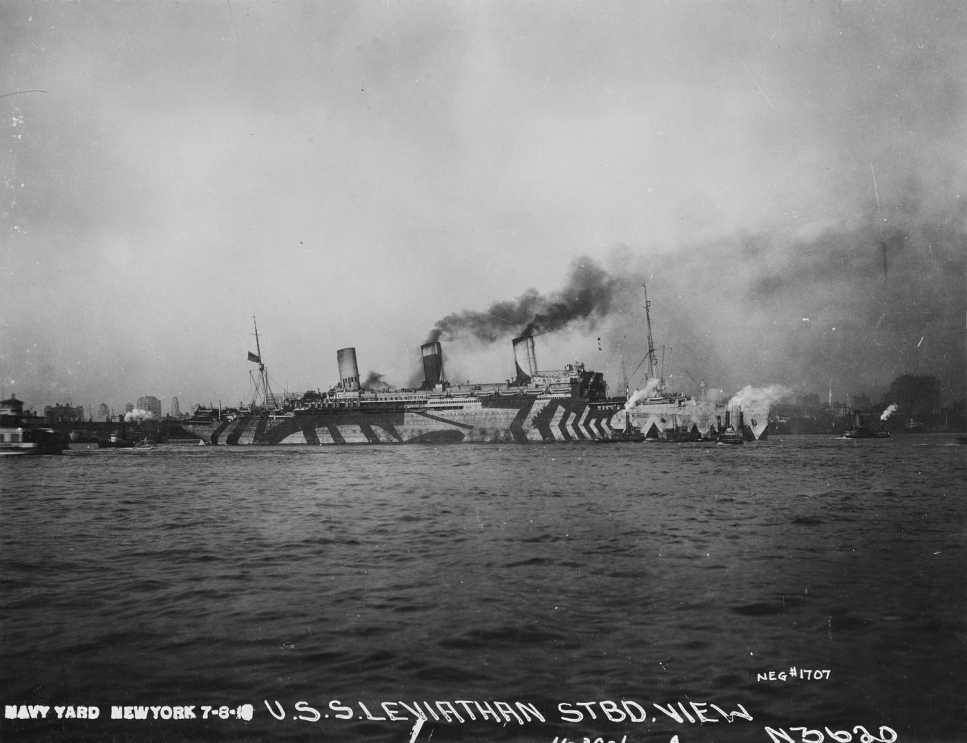 Black-and-white photograph of a huge four-funnel ship painted in bold zigzag camouflage stripes, with tugs alongside in a harbor