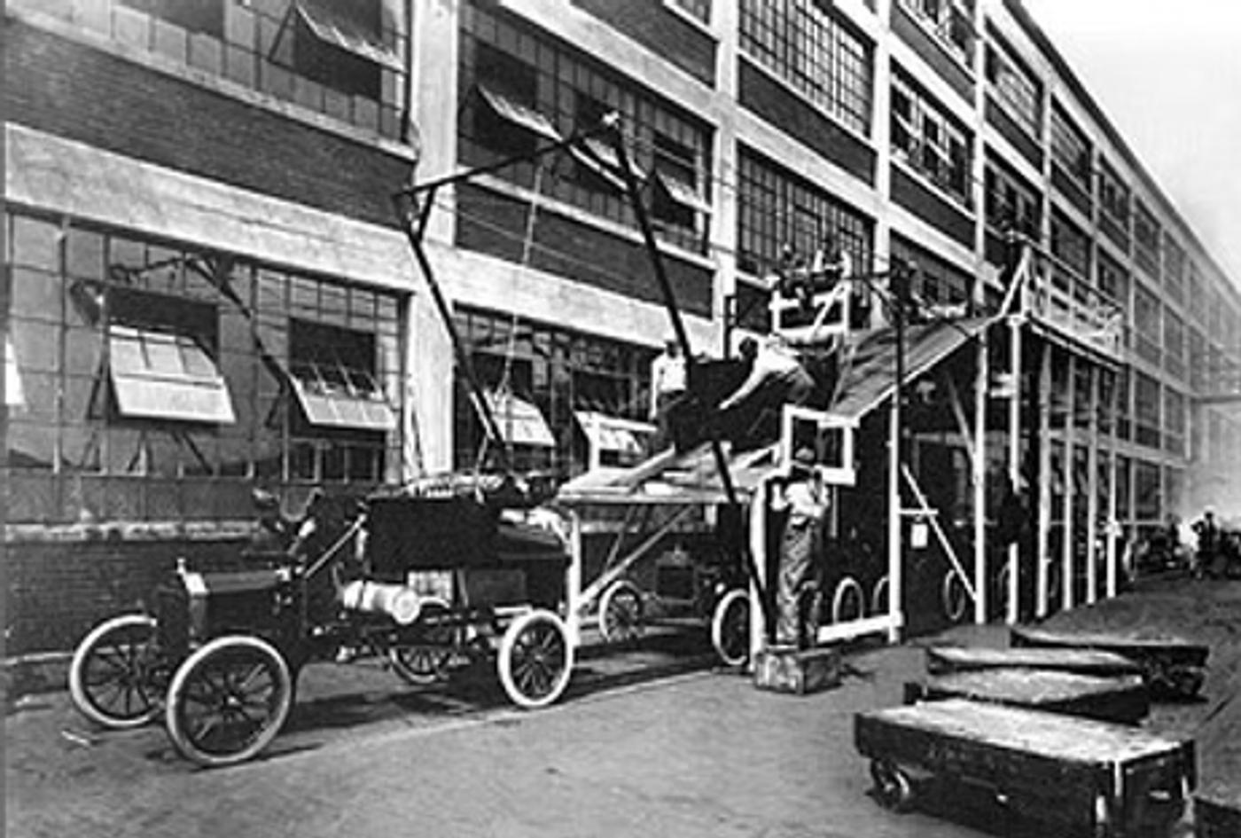 Early automotive assembly line with workers, car frames on stands, and vintage vehicles outside industrial factory building, circa 1913-1920.