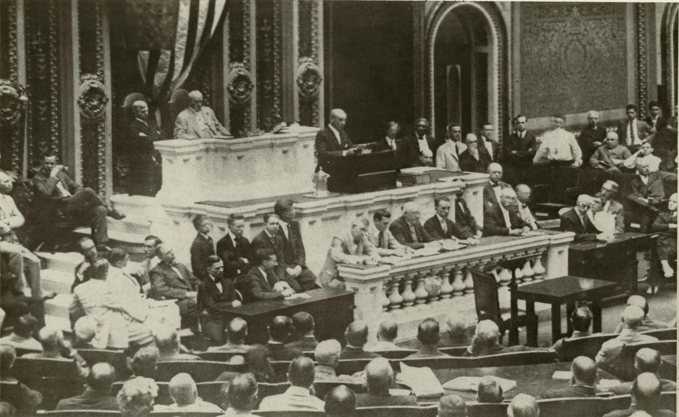 Black-and-white photograph of a man reading a speech at a marble rostrum in the crowded House chamber, with members of Congress seated in rows before him