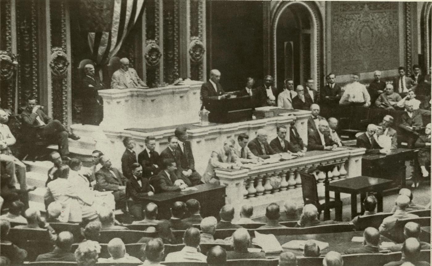 Black-and-white photograph of a man reading a speech at a marble rostrum in the crowded House chamber, with members of Congress seated in rows before him