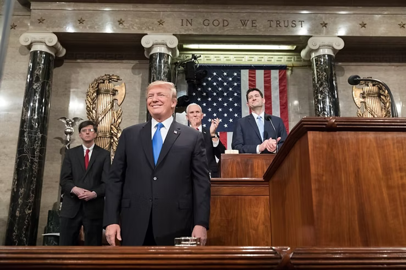 Man in dark suit with blue tie stands at wooden podium in ornate Capitol chamber with American flag and "In God We Trust" inscription visible.