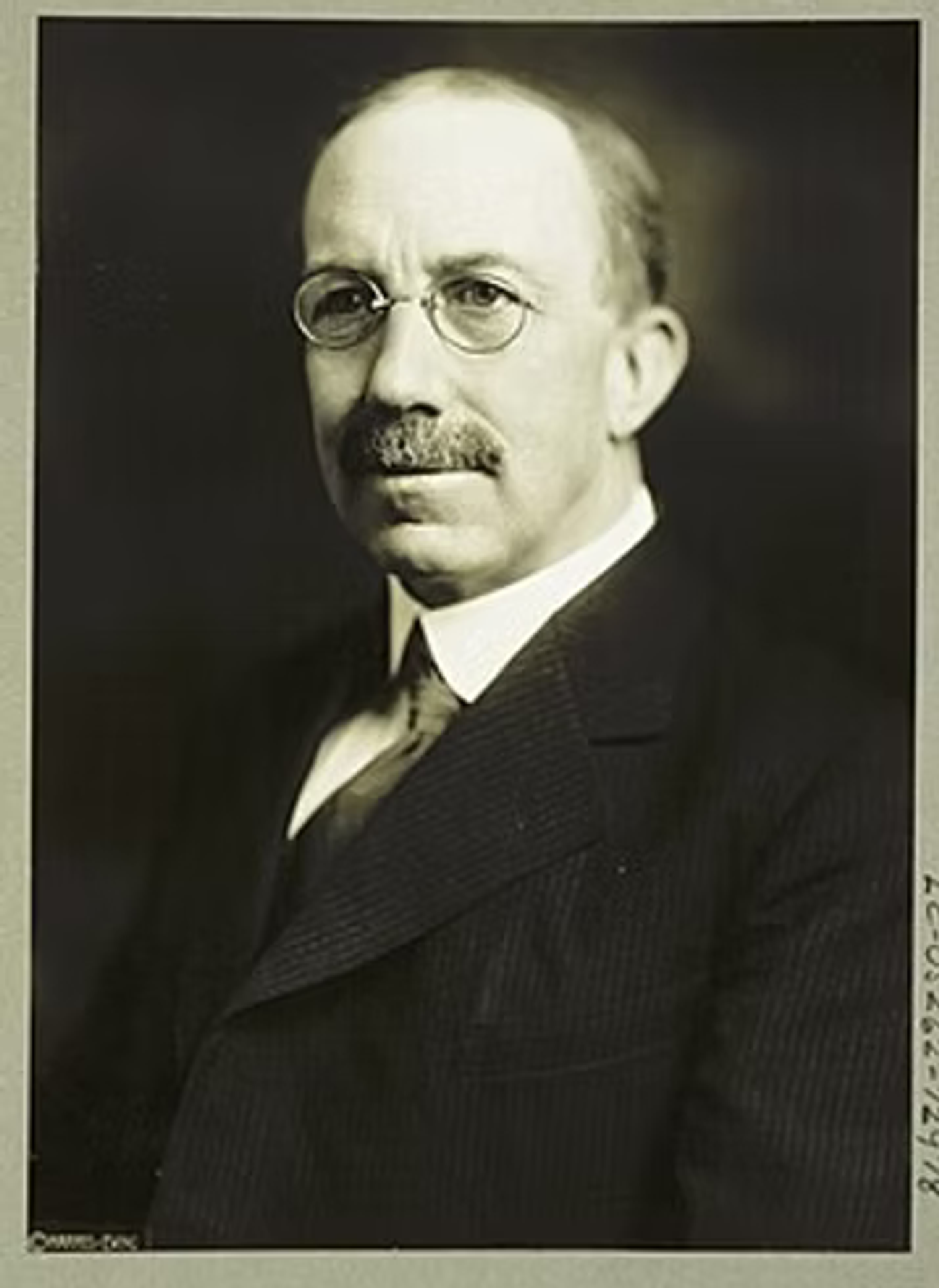 Formal black and white portrait of a bald man with a mustache wearing round glasses and a dark suit with white shirt and tie.