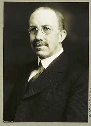 Formal black and white portrait of a bald man with a mustache wearing round glasses and a dark suit with white shirt and tie.