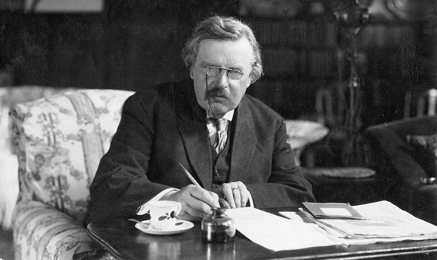 Elderly man with glasses and mustache writing at desk in formal suit, circa 1913-1920.