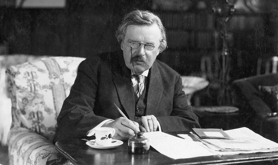 Elderly man with glasses and mustache writing at desk in formal suit, circa 1913-1920.