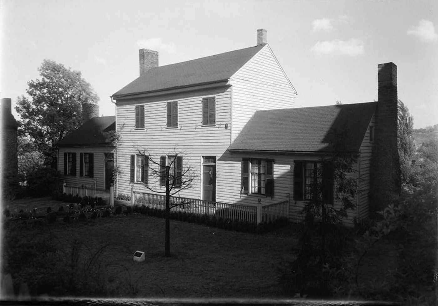 Black-and-white photograph of a white two-story frame house with dark shutters and brick chimneys, flanked by lower wings, behind a picket fence and garden