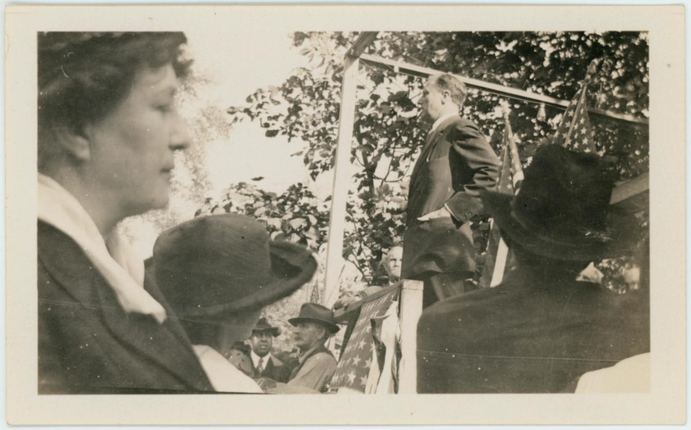 Black-and-white photograph of a young man in a dark coat speaking from a raised platform under trees, seen past women in wide-brimmed hats in the foreground