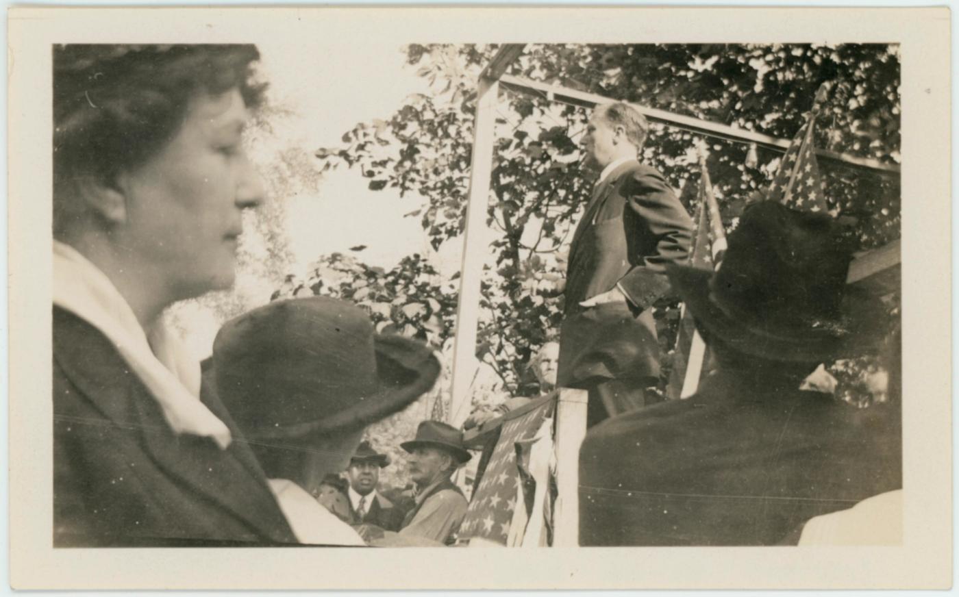 Black-and-white photograph of a young man in a dark coat speaking from a raised platform under trees, seen past women in wide-brimmed hats in the foreground