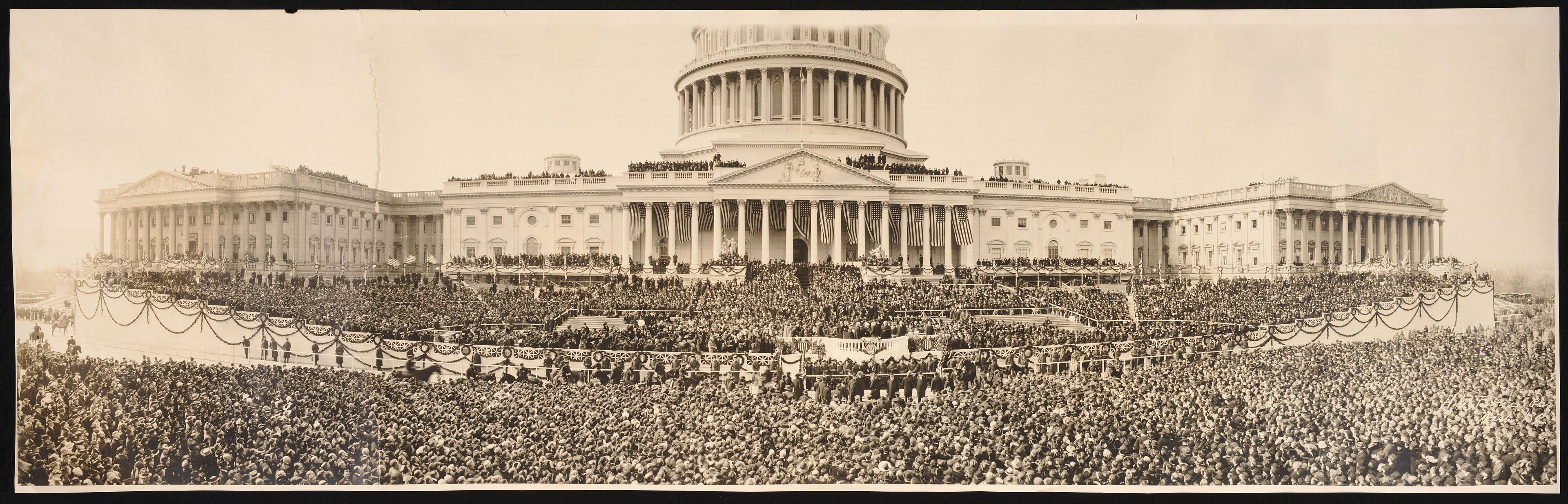 Sepia panoramic photograph of a vast crowd filling the plaza in front of the U.S. Capitol, its dome and wings hung with flags and bunting for a presidential inauguration