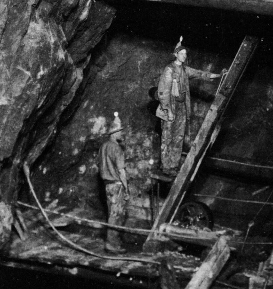 Flash-lit photograph deep in a mine: two miners with candles fixed to their hats stand on timber platforms against a rough rock wall, one riding the stepped beam of a man engine