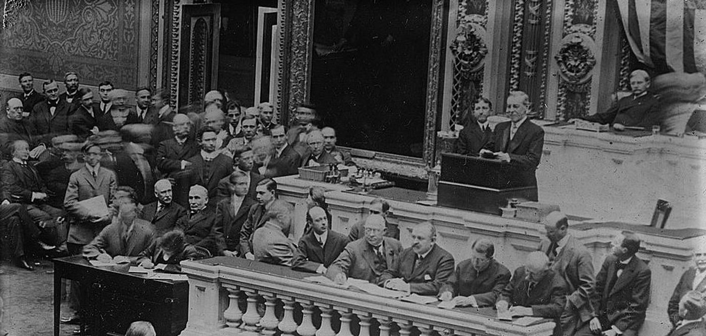 Woodrow Wilson addressing congress. Black and white photograph of a formal legislative session in an ornate chamber with officials seated at a raised desk and spectators in the gallery.