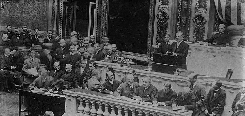 Woodrow Wilson addressing congress. Black and white photograph of a formal legislative session in an ornate chamber with officials seated at a raised desk and spectators in the gallery.
