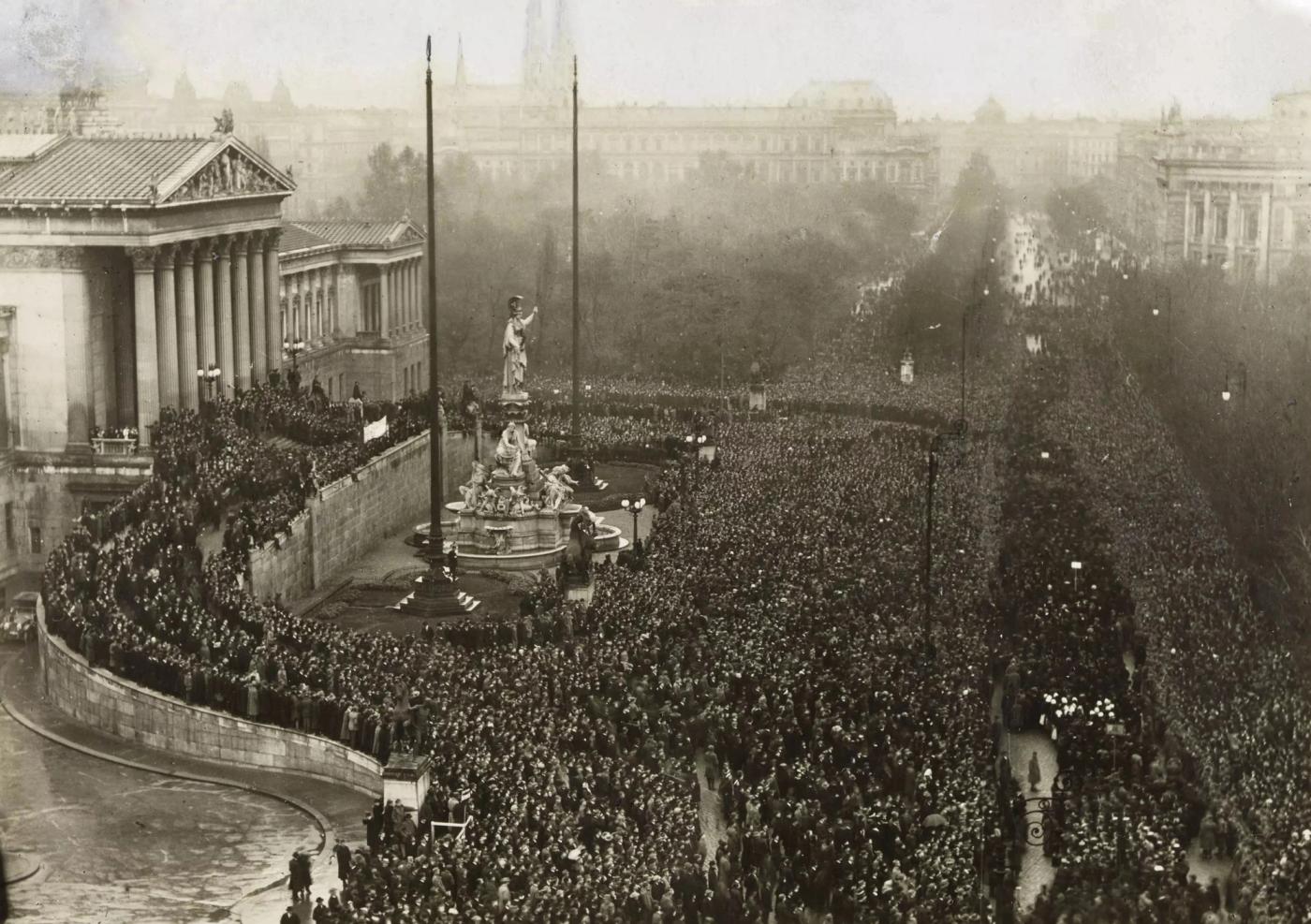 Elevated view of a vast crowd filling the Ringstraße in front of a columned classical parliament building, with flagpoles and a monumental fountain in the foreground