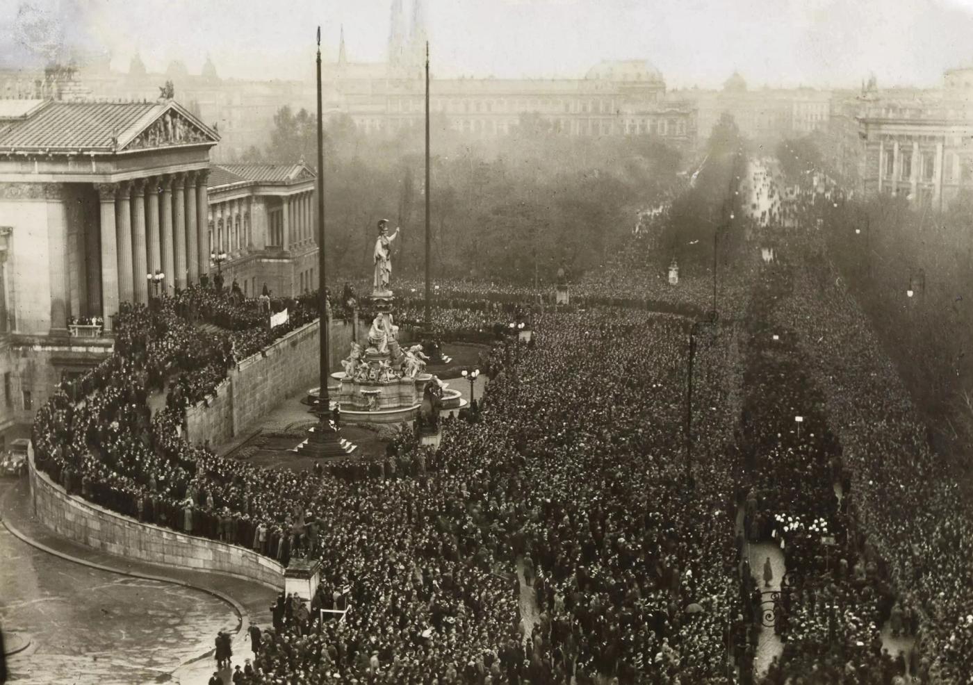 Elevated view of a vast crowd filling the Ringstraße in front of a columned classical parliament building, with flagpoles and a monumental fountain in the foreground
