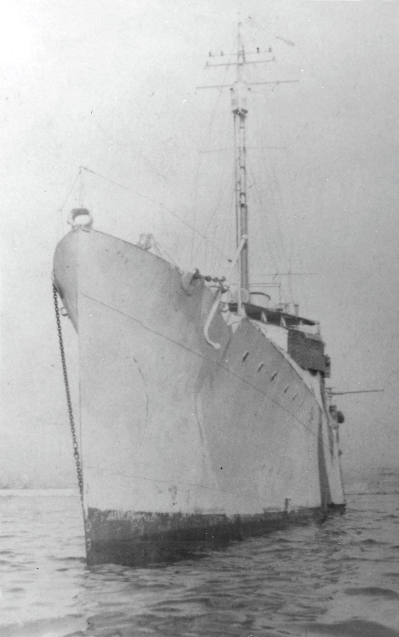 Black-and-white photograph of the bow of a narrow destroyer seen head-on, with a tall mast, riding on calm water