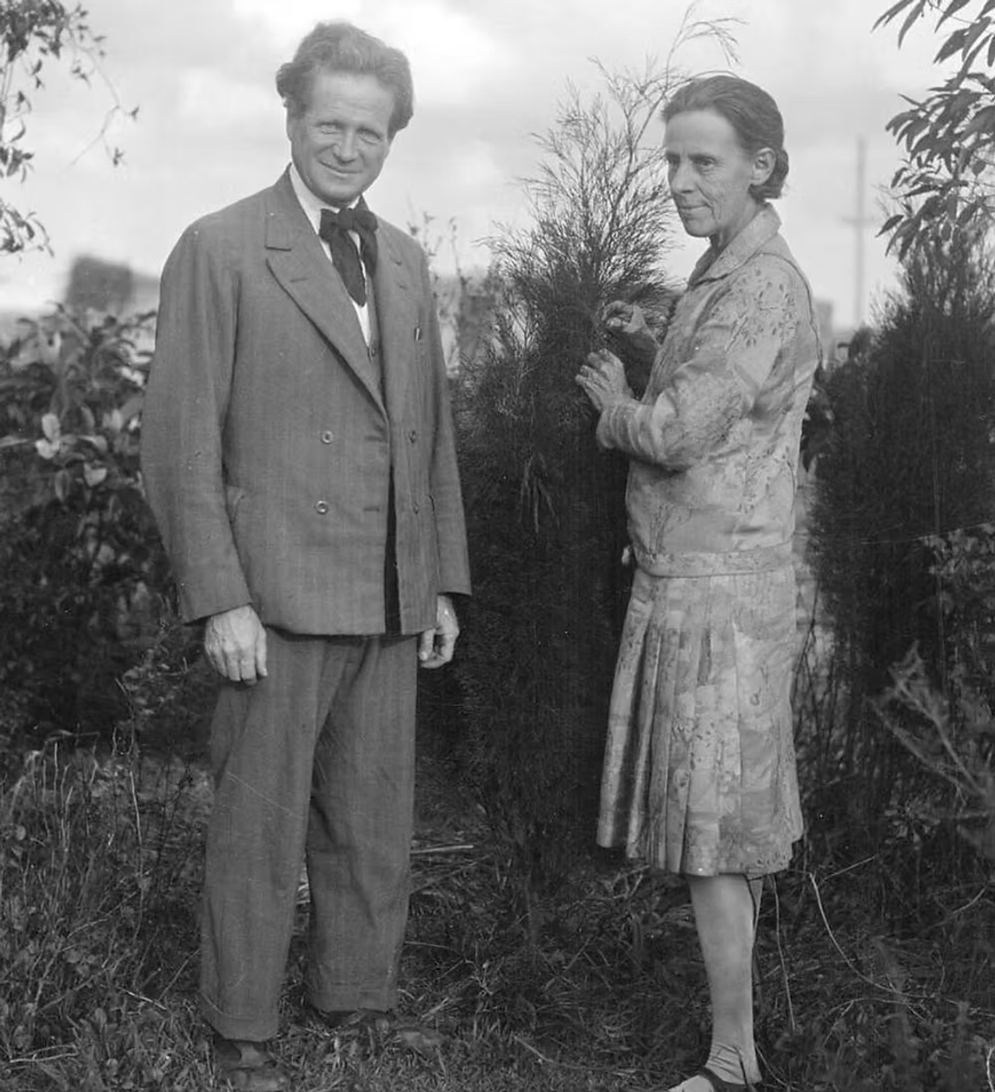 Two people in 1920s-style clothing standing in a garden holding a potted ornamental grass plant.