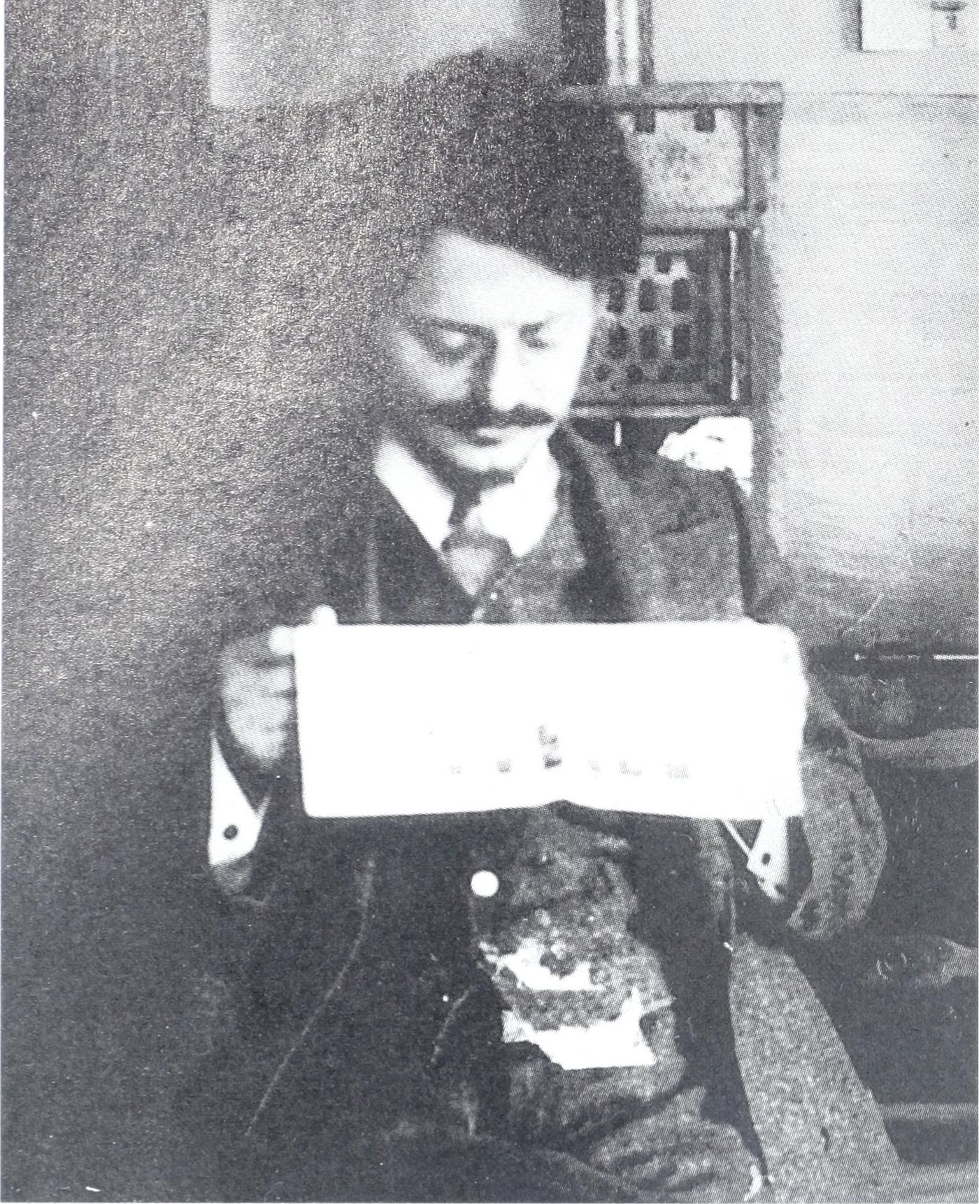 Black-and-white photograph of a young man with wavy hair and a moustache, in a jacket and tie, holding open a newspaper