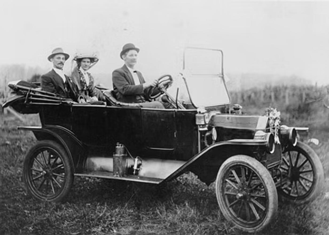 Early 1900s automobile with three passengers in formal attire driving on rural road with forest background.