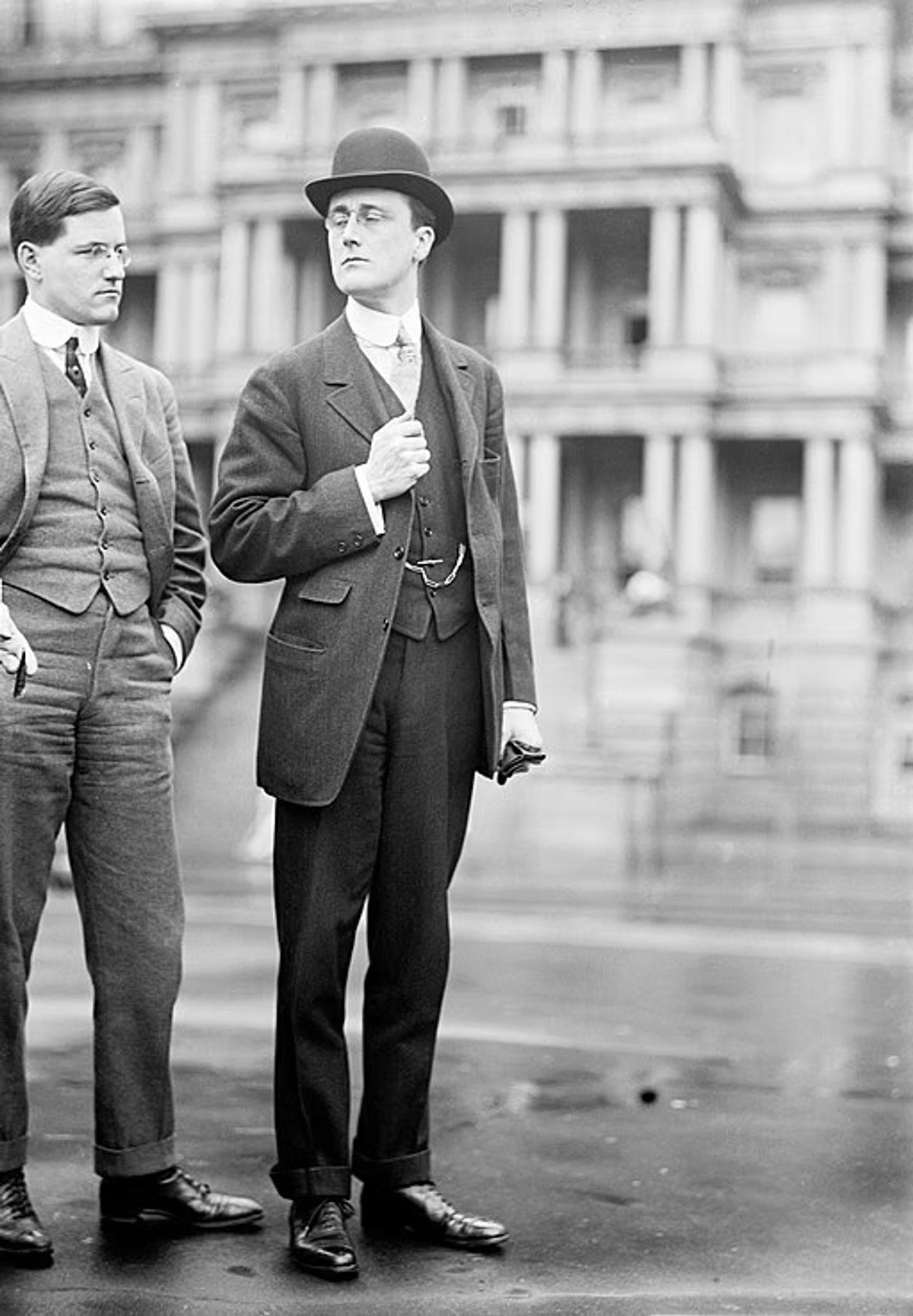 Two men in suits and bowler hats stand outside a classical government building, circa early 1900s.