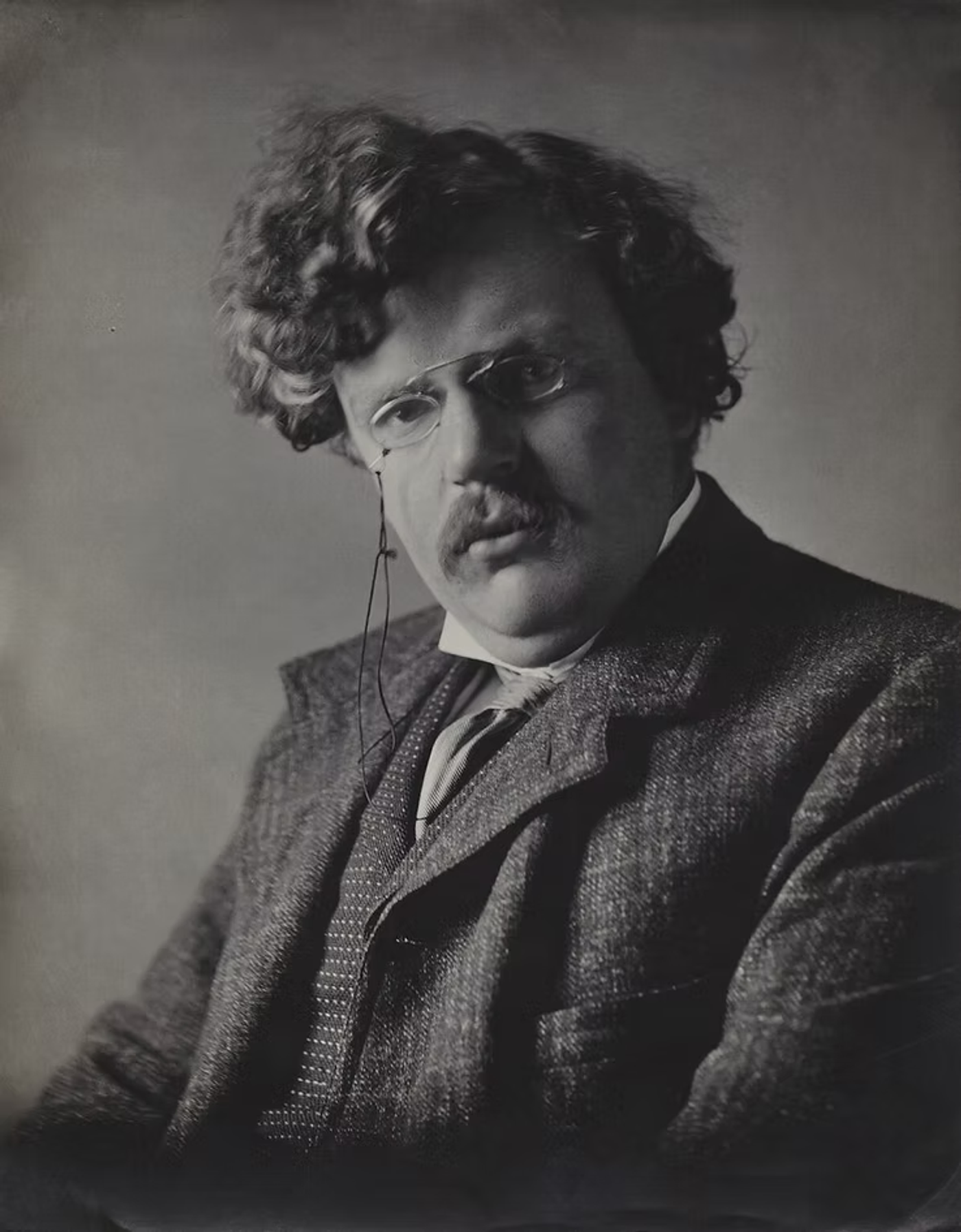 Black and white portrait of a man with wavy hair and mustache wearing glasses and a tweed jacket, circa 1913-1920.