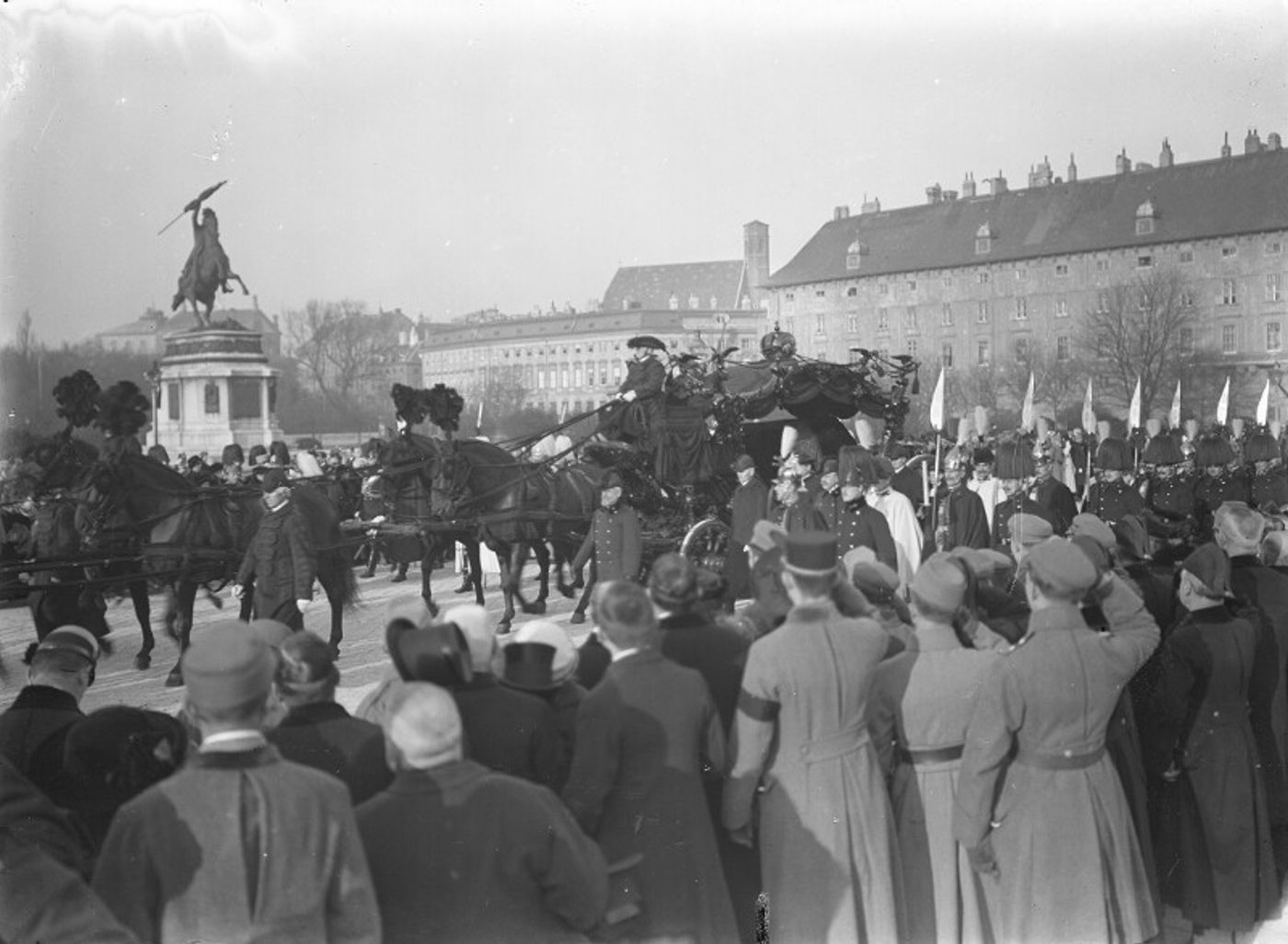 Black-and-white photograph of a horse-drawn funeral procession passing a crowd of onlookers in a large square, with an equestrian statue and a long palace wing behind