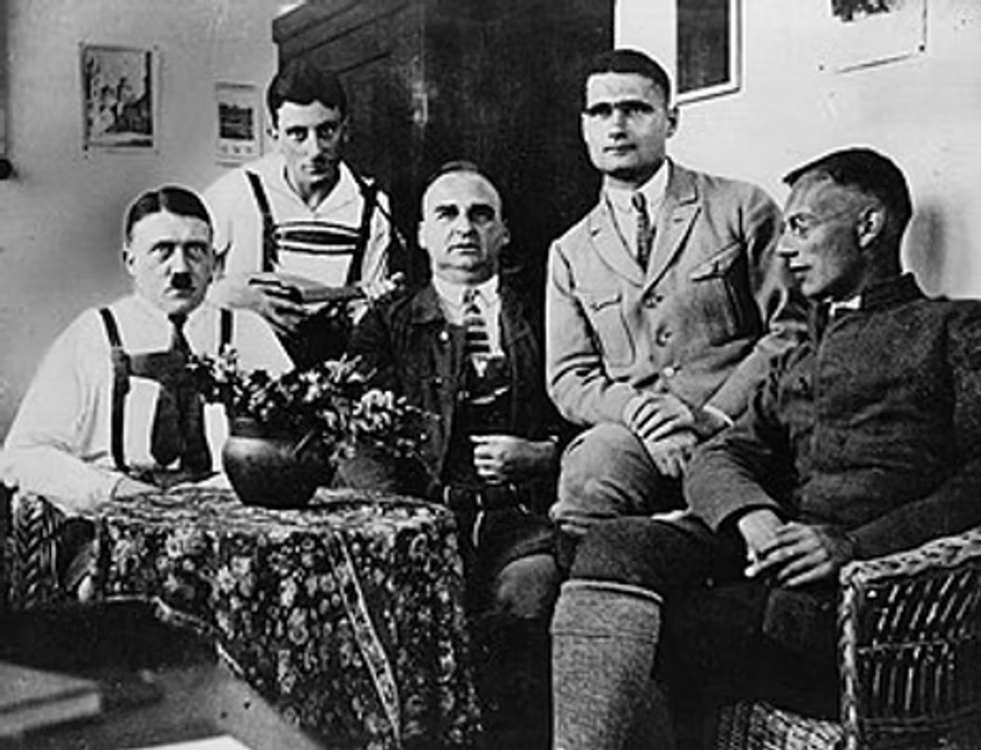 Five men in 1920s-era clothing seated and standing in an interior room with a potted plant on a table.