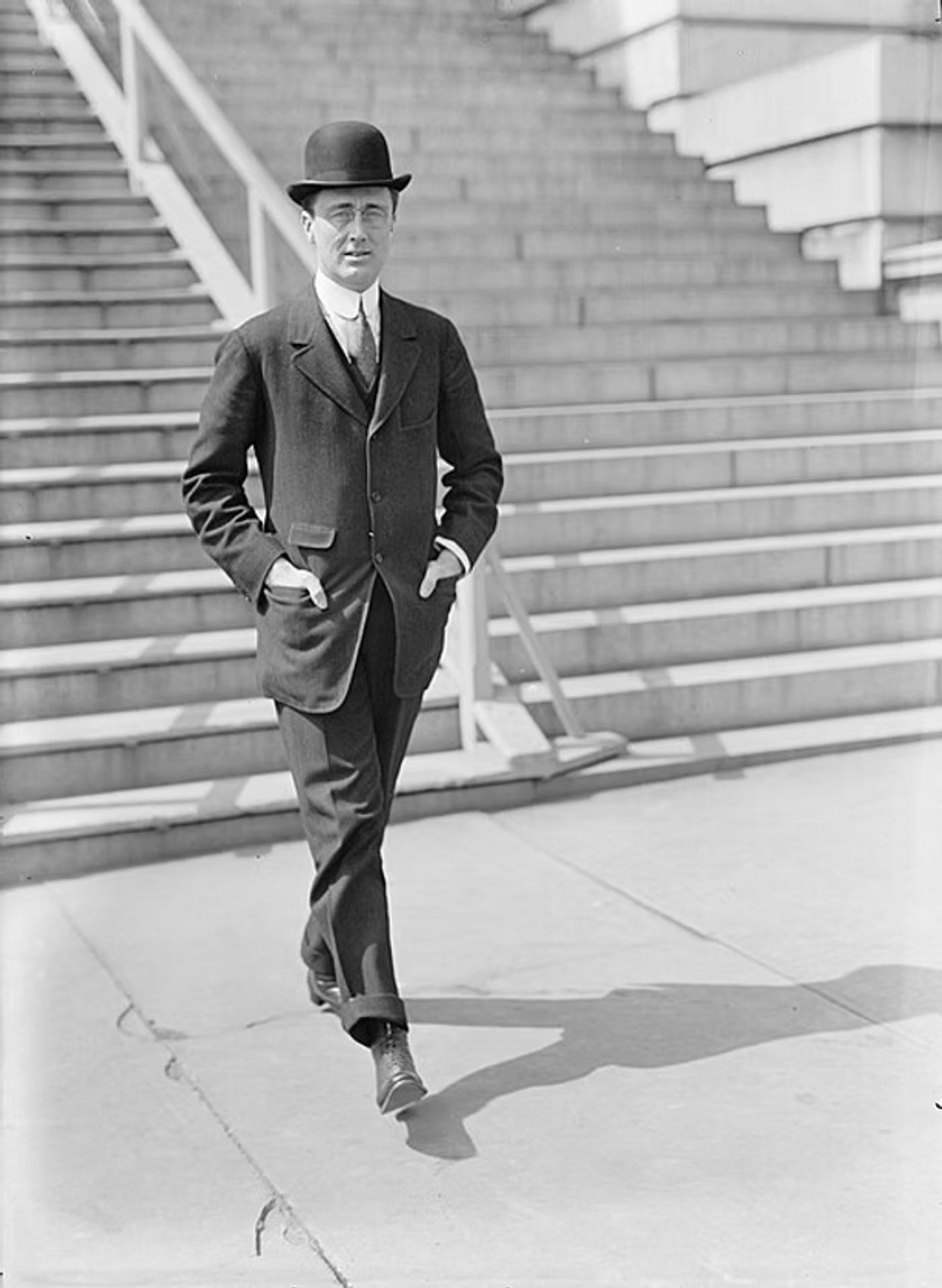 Man in bowler hat and dark suit walking on stone steps near brick building, early 20th century.