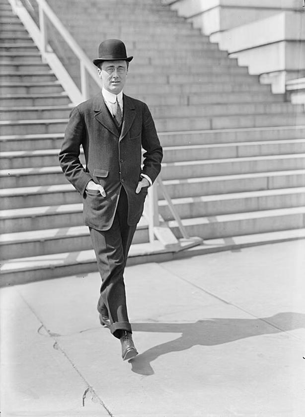 Man in bowler hat and dark suit walking on stone steps near brick building, early 20th century.