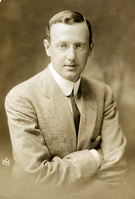 Man in suit and glasses, early 20th century portrait photograph with neutral background.