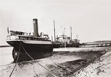 Steamship moored at sandy shore with industrial facility and multiple smokestacks visible in background, circa early 20th century.