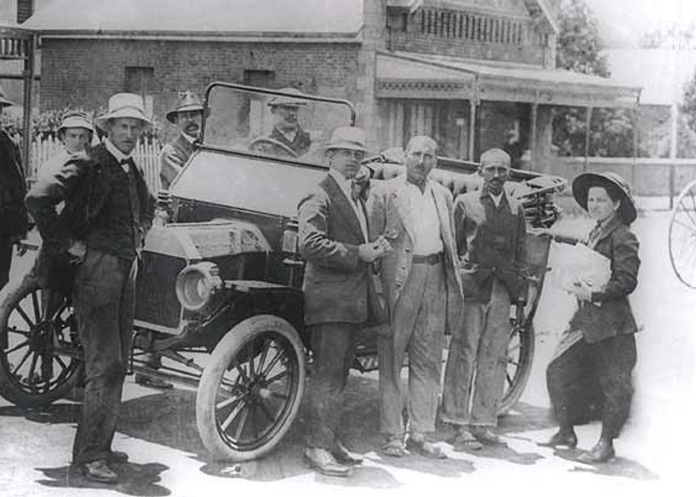 Black and white historical photograph of Gandhi with group of men and women around early automobile and bicycles in front of houses.