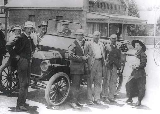 Black and white historical photograph of Gandhi with group of men and women around early automobile and bicycles in front of houses.