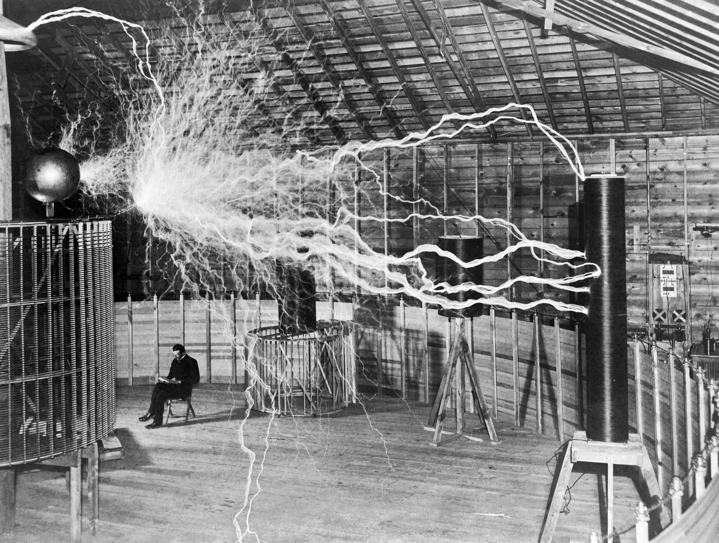 Black-and-white photograph of an electrical laboratory where massive arcs of electricity surge between large coils and equipment. A man sits calmly in a chair near the center of the room, reading while bright lightning-like discharges fill the space around him. The image, one of the most famous photographs associated with Nikola Tesla, demonstrates high-voltage experiments conducted at his Colorado Springs laboratory in 1899.