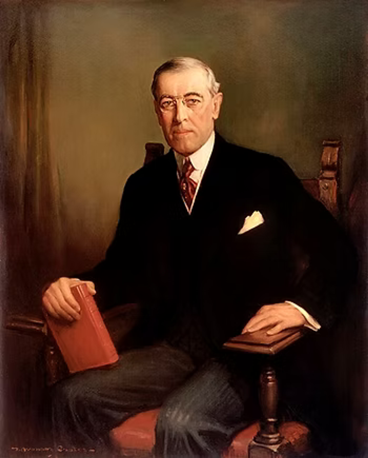 Portrait of a man in formal black suit with red tie, holding a red book, seated in a chair against a neutral background.