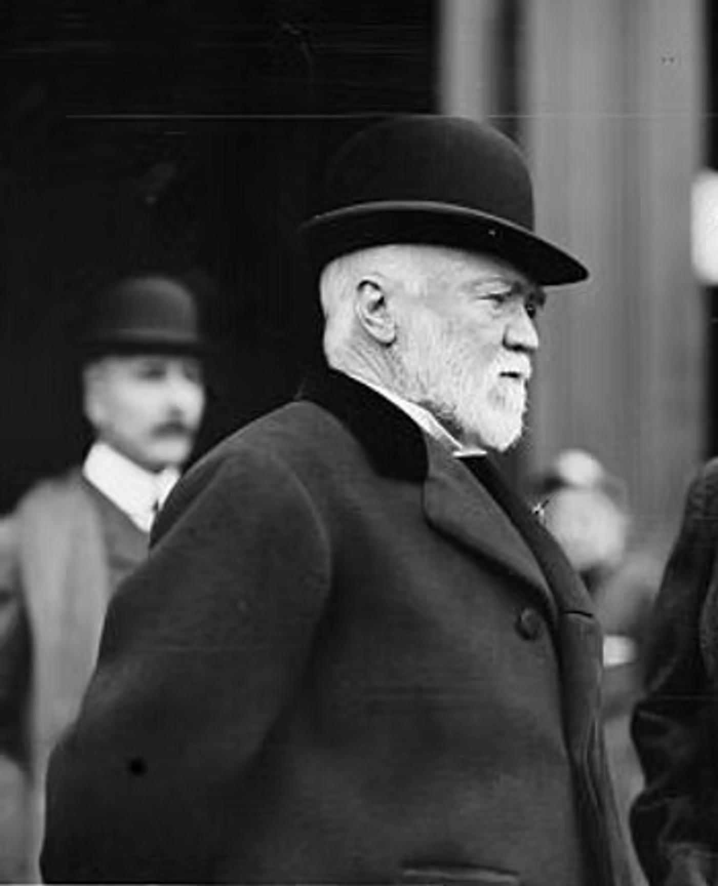 Andrew Carnegie with white beard wearing dark coat and bowler hat, photographed in profile with other figures blurred in background.