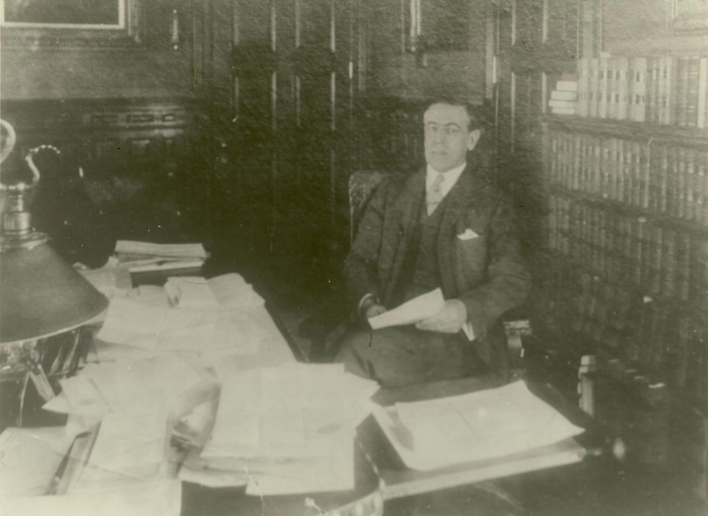 Faded photograph of a man in a three-piece suit seated beside a desk piled with papers, holding a document, with tall bookcases behind him