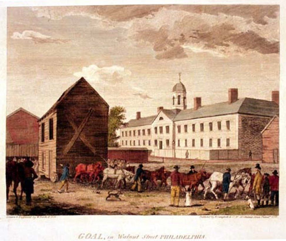 Hand-coloured engraving of a long two-storey stone prison with a central cupola standing behind a wall, with teams of harnessed horses, drovers and onlookers crowding the open ground in front of it