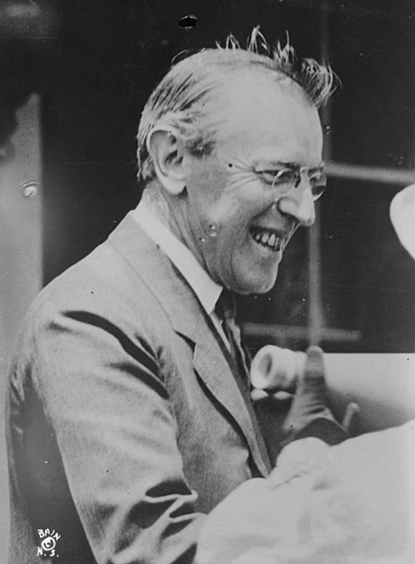 Woodrow Wilson wearing glasses and suit jacket, smiling while talking.