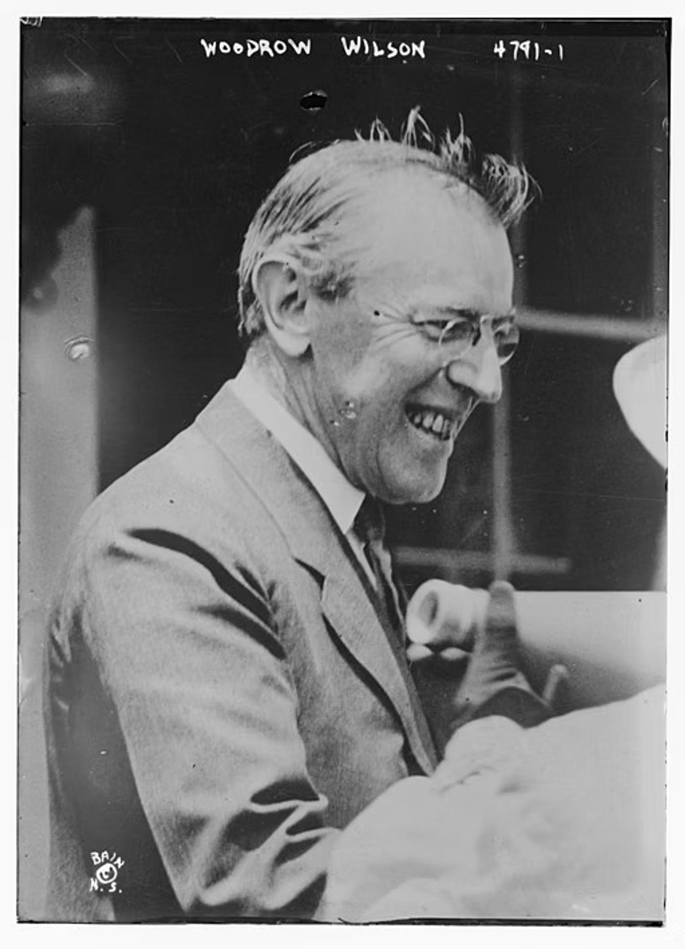 Woodrow Wilson wearing glasses and suit jacket, smiling while talking.