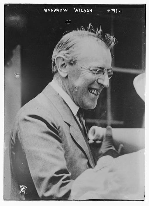 Woodrow Wilson wearing glasses and suit jacket, smiling while talking.