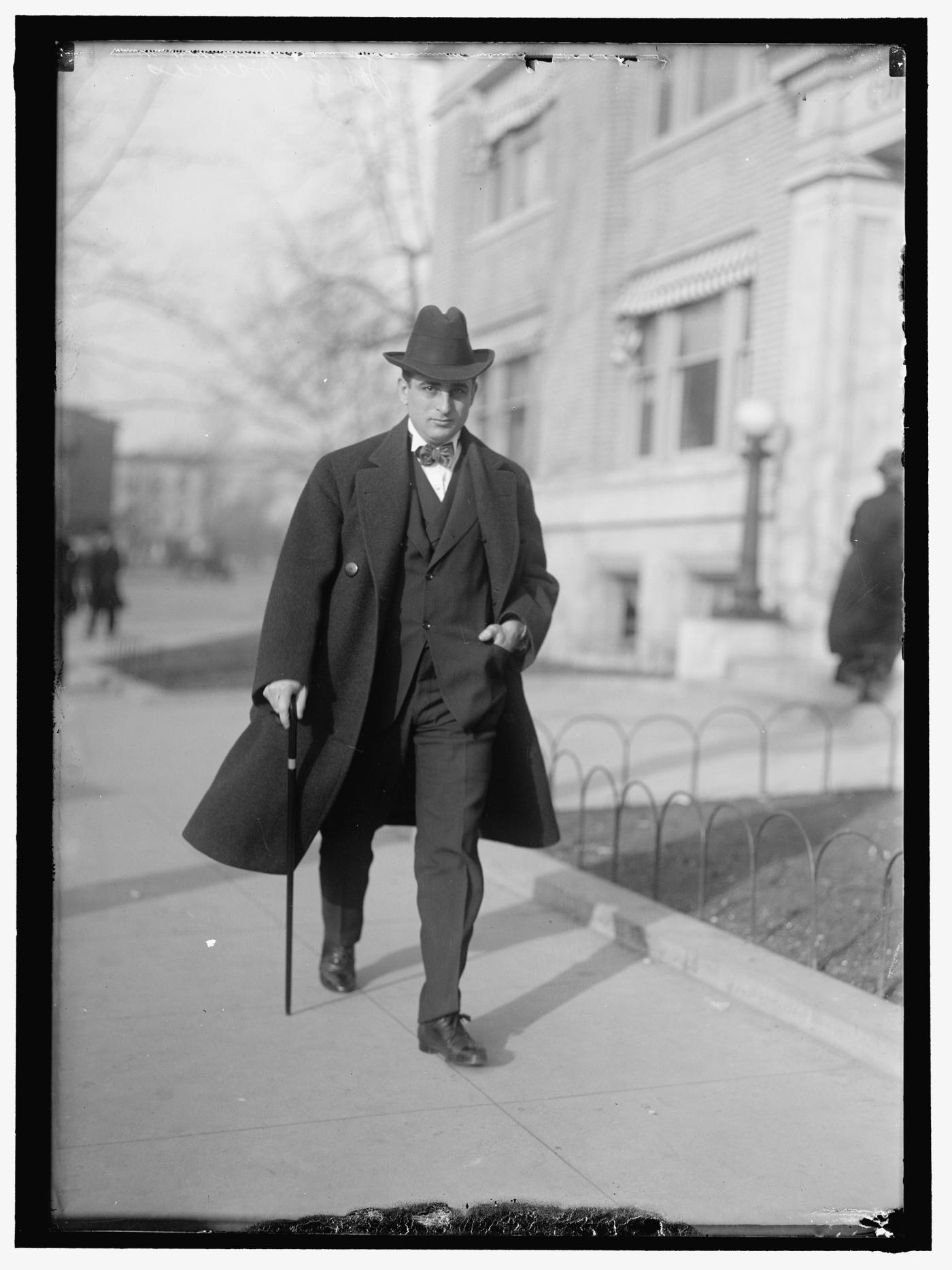 Black-and-white photograph of a man in a dark overcoat and hat walking along a sidewalk outside a stone building