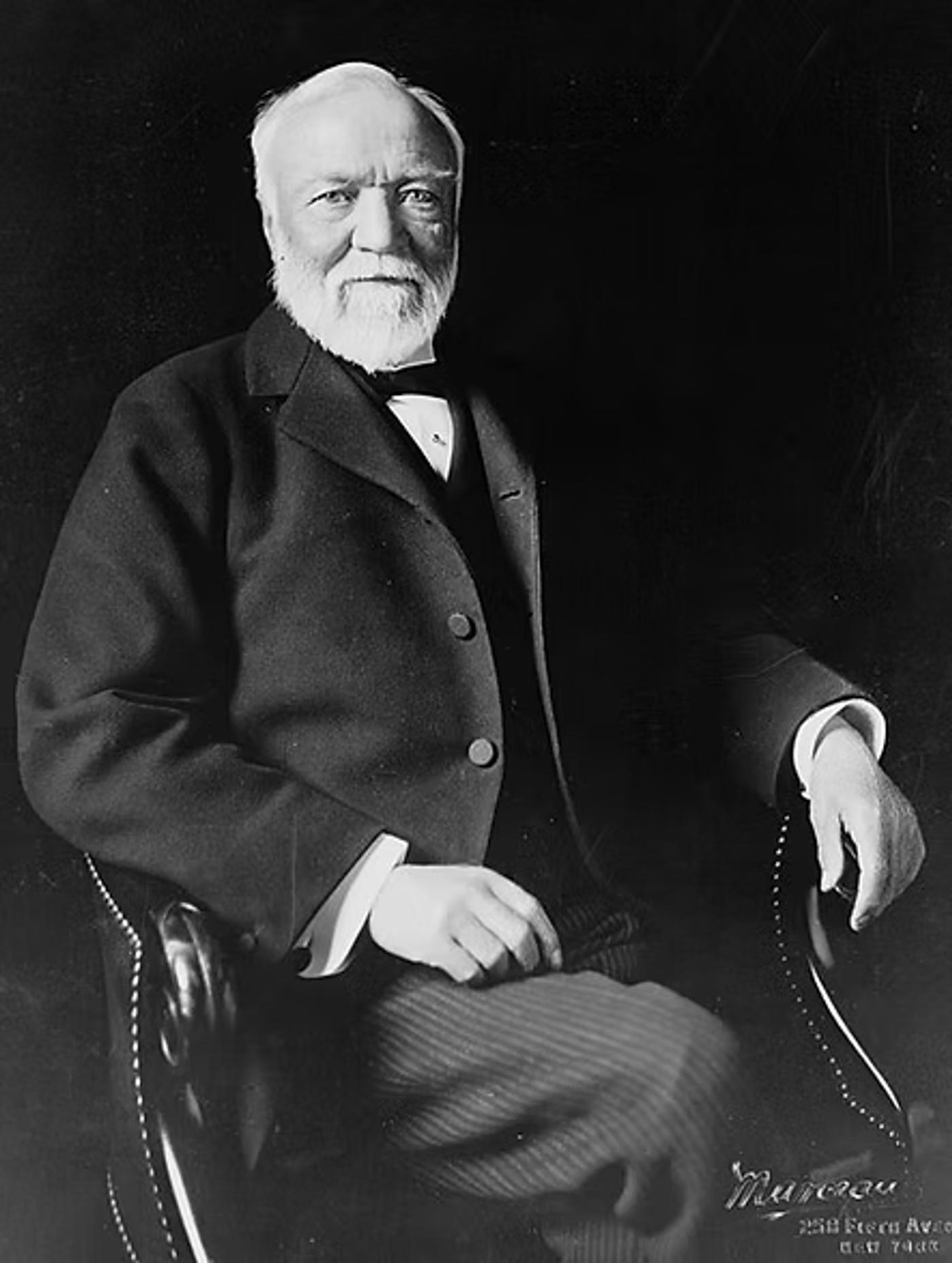 Andrew Carnegie with white beard in formal dark suit and bow tie, seated against black background, circa early 1900s.