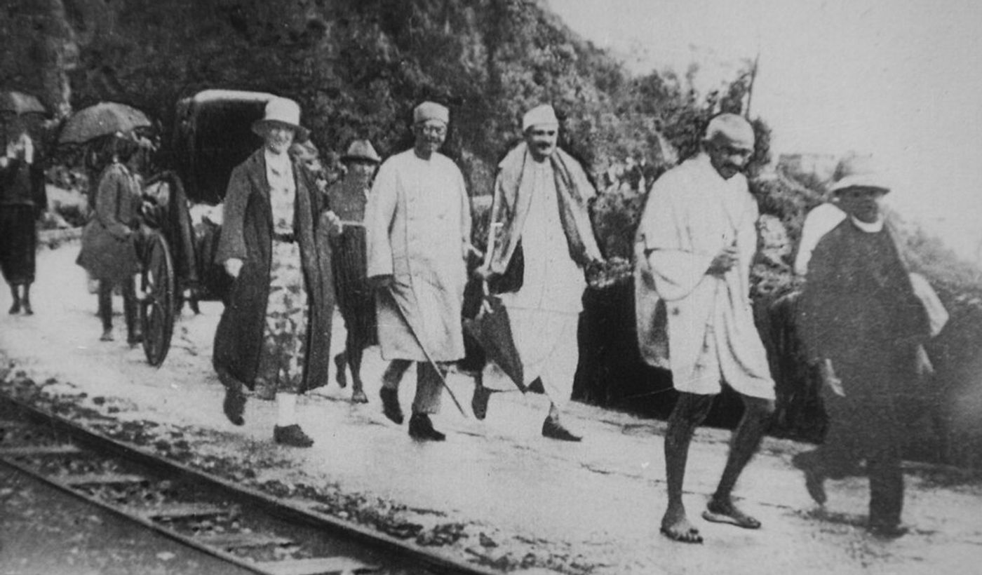 Gandhi walking with Deshbandu C.R. Das on a tree-lined path, surrounded by followers in traditional Indian attire.
