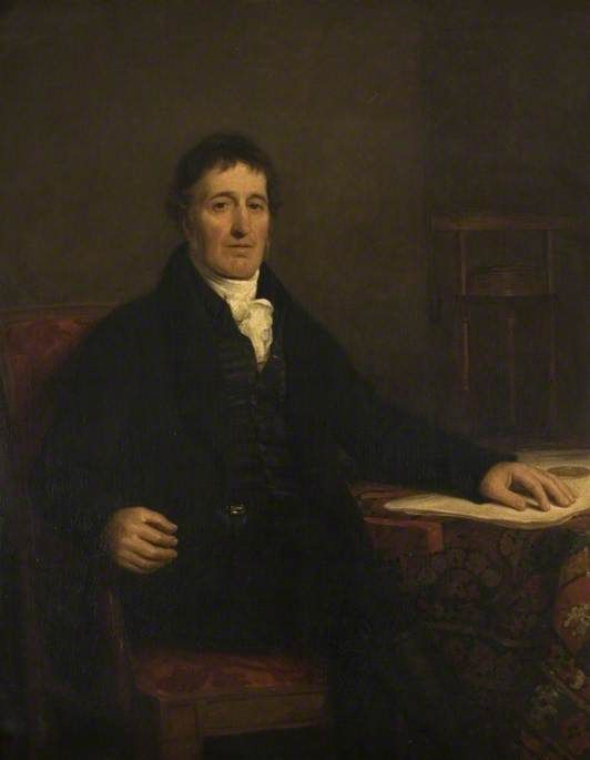 Portrait of William Murdoch, seated at a table with papers, in a dark coat