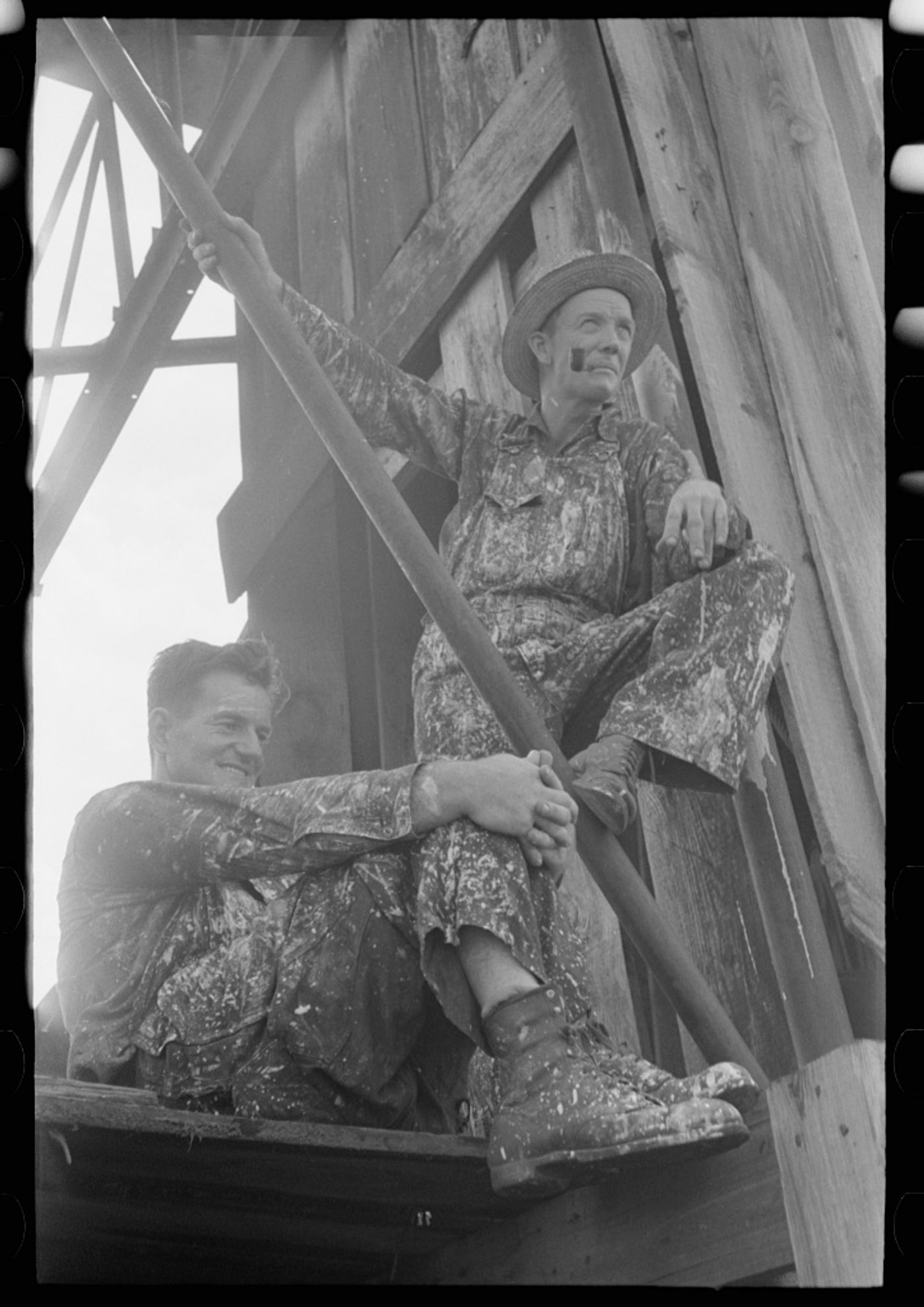 Two workers in patterned work clothes sit among oil derrick wooden beams at an Oklahoma oil field facility.