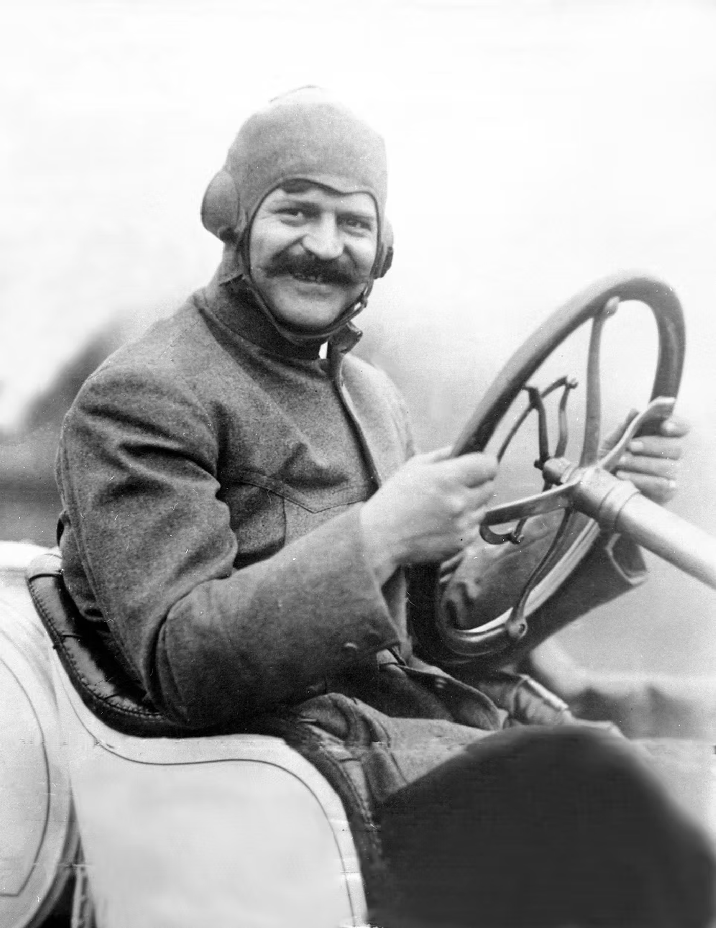 Man with mustache and aviator cap at steering wheel of early automobile, circa 1910s-1920s.