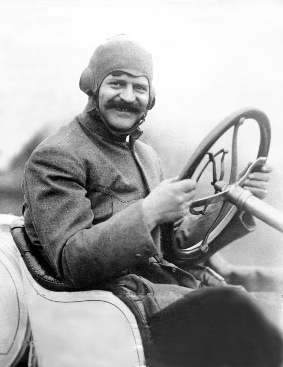 Man with mustache and aviator cap at steering wheel of early automobile, circa 1910s-1920s.