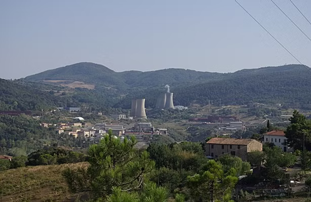 Industrial cooling towers in a forested valley with traditional European buildings in the foreground and mountains in the background.