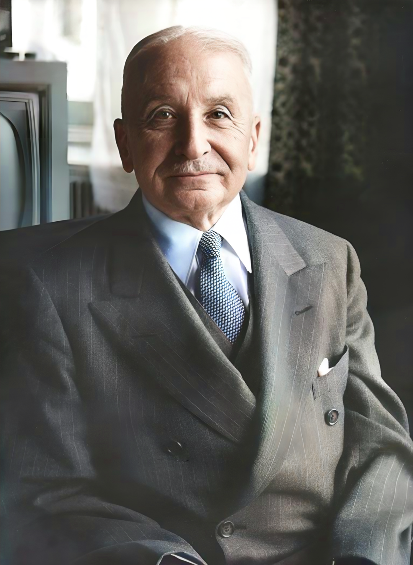 Elderly man in gray pinstriped suit and blue tie, seated indoors with window and ivy visible in background.