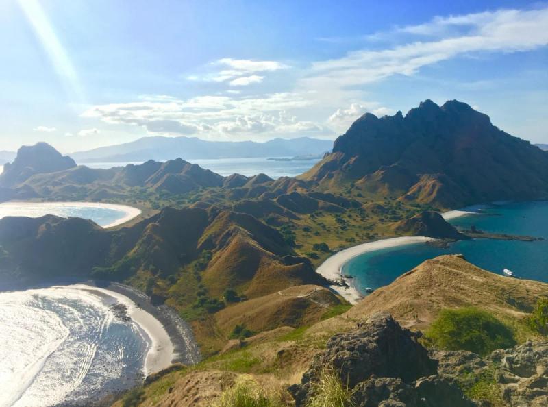 Pemandangan panorama Pulau Padar di Taman Nasional Komodo: bukit curam, teluk biru toska dengan pantai putih dan abu-abu di bawah langit cerah